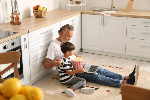 Little boy and his grandfather with tasty popcorn using laptop in kitchen - Фото, изображение