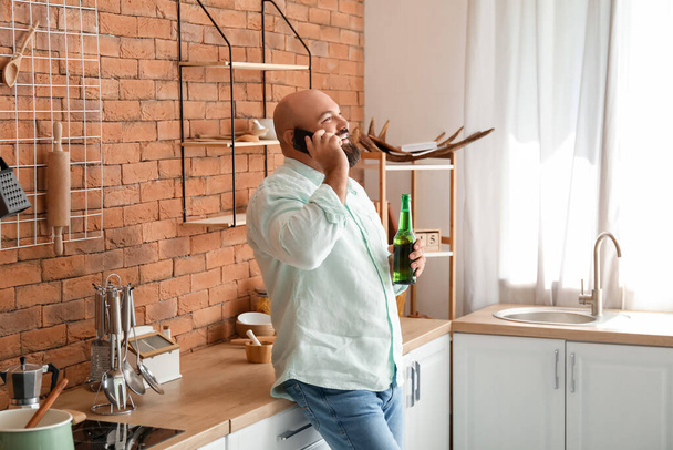 Bald man drinking beer and talking by phone in kitchen - Zdjęcie, obraz