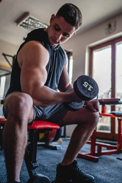 One man young adult caucasian male bodybuilder training arms bicep flexing muscles with dumbbell while sitting in the gym wearing shirt real people copy space front view - Foto, Imagem