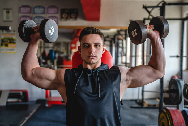 One man young adult caucasian male bodybuilder training shoulder press deltoid muscles with dumbbell while sitting in the gym wearing shirt real people copy space front view - Фото, зображення