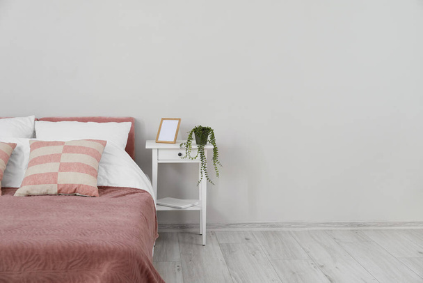 Comfortabel bed, nachtkastje met houseplant en blanco frame bij lichte wand - Foto, afbeelding