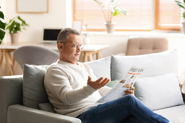 Upset senior man in eyeglasses reading newspaper on sofa at home - Foto, Imagen