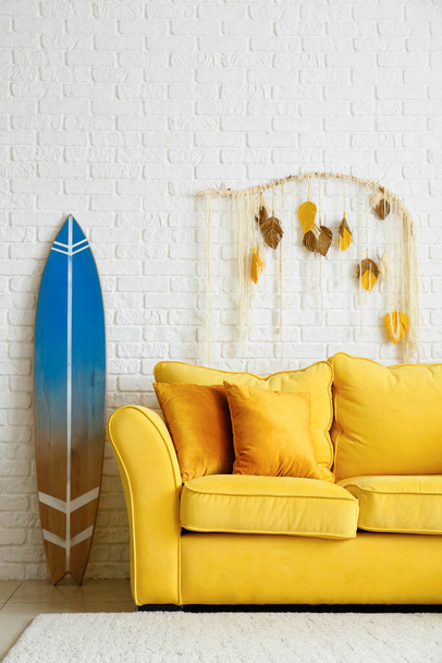 Удобный диван и доска для серфинга возле стены из белого кирпича - Фото, изображение