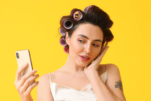 Beautiful young woman in hair curlers taking selfie on yellow background - Фото, изображение