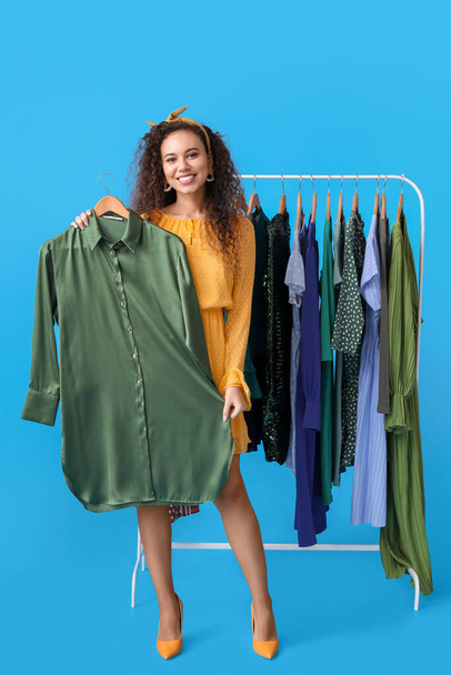 Beautiful African-American girl near rack with clothes for high school prom on blue background - Фото, зображення