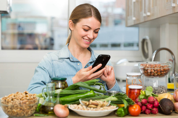 Portrait of glad woman looking at mobile phone while making dish on kitchen at home - Fotoğraf, Görsel