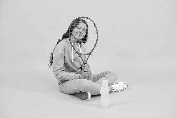 glückliches Kind sitzt in Sportbekleidung mit Squash-Schläger und Wasserflasche auf gelbem Hintergrund, Kindheit - Foto, Bild