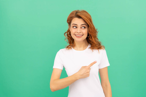 χαμογελαστή κοκκινομάλλα γυναίκα με σγουρά μαλλιά δείχνοντας το δάχτυλο σε μπλε φόντο, αντίγραφο χώρου, διαφήμιση - Φωτογραφία, εικόνα