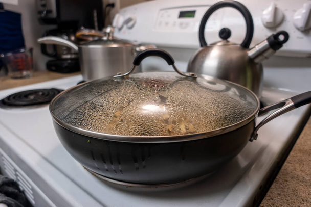 Enfoque selectivo en ángulo de una gran sartén cubierta que cocina la cena a fuego lento en una estufa blanca, condensación y vapor que se forma en el interior - Foto, imagen