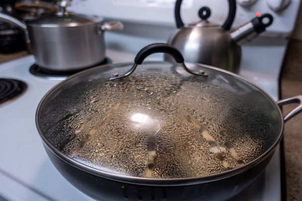 Angled, szelektív fókusz egy nagy, fedett serpenyő főzés vacsora egy pároló egy fehér tűzhely tetején, páralecsapódás és gőz képződés belül - Fotó, kép