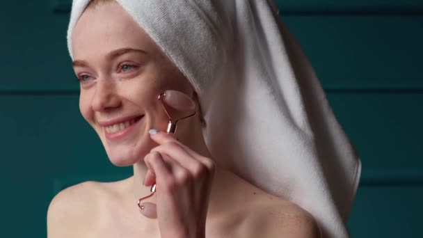 Mosolygó kaukázusi nő fürdőruhában masszírozza az arcát jáde arcgörgővel, hogy ellazítsa az izmokat. Megfiatalító kezelés. Bőrgyógyászat, kozmetika. Orvosi - Felvétel, videó