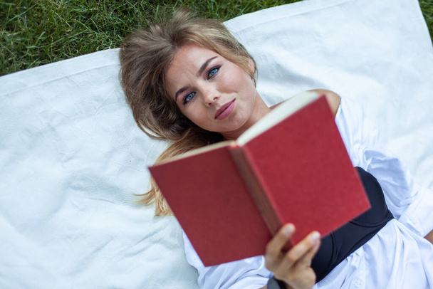 Linda mujer atractiva lectura libro mientras está acostado en la manta en la hierba  - Foto, Imagen