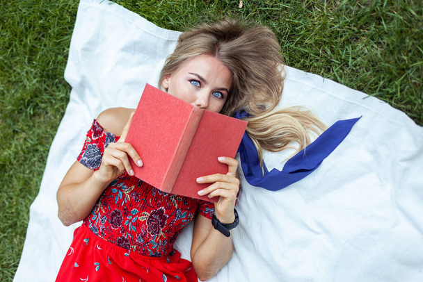 Hübsche Frau im roten Kleid mit einem Buch auf einem Karo im Park. Freizeitgestaltung im Freien - Foto, Bild