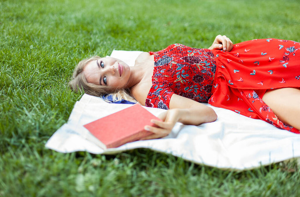 Красивая женщина в красном платье с книгой о клетчатом в парке. Открытый отдых на природе - Фото, изображение