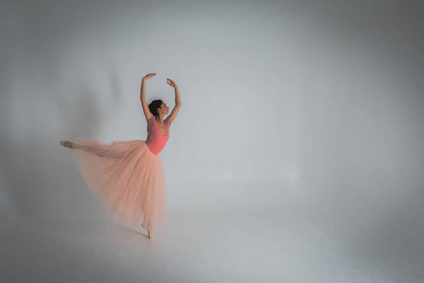 νέα όμορφη, εύθραυστη, όμορφη μπαλαρίνα χορεύει σε ένα μακρύ ανοιχτό ροζ φόρεμα με τούλι σε ένα ομοιόμορφο φόντο, χαμηλό κλειδί. Μπαλέτο, χορός, χορευτής - Φωτογραφία, εικόνα