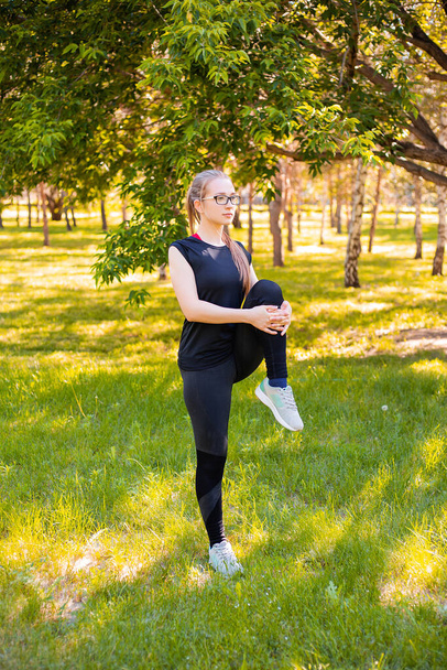 A girl in a sports uniform does a warm-up and stretches her legs while standing in a summer park. Outdoor sports and fitness. - Foto, afbeelding