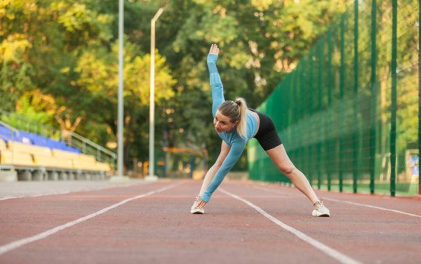 Sportowa kobieta robi rozgrzewkę przed treningiem na stadionie. Zdrowy styl życia - Zdjęcie, obraz