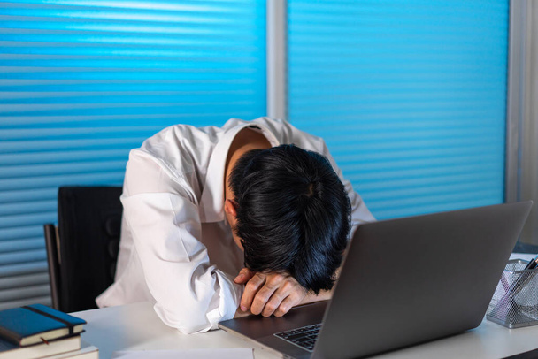 Der Mann, der spät im Büro arbeitet, den Kopf auf dem Schreibtisch liegend, während er vor dem Laptop arbeitet. - Foto, Bild