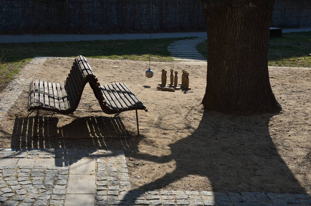 černá kovová křesla a kulaté stoly v kavárně na dlážděném náměstí s pískem pokrytým zhutněným žlutým štěrkem pod starým stromem. Ruské dřevěné kuželky a koule na řetězu - Fotografie, Obrázek