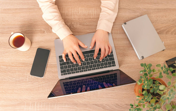 Nahoře je fotografie pracovního stolu ženy, která píše do laptopu. Rostlina, telefon, pohár a deník jsou také na stole. - Fotografie, Obrázek