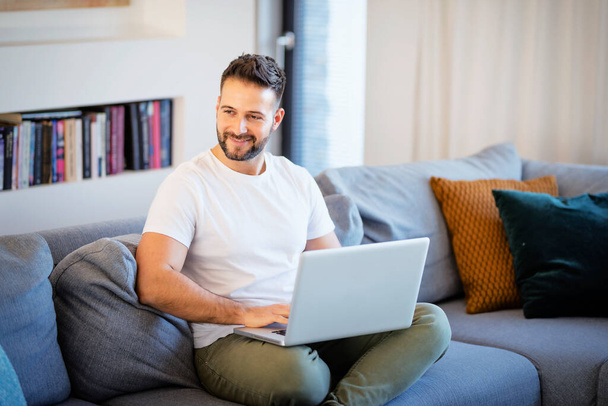 Handsome man using laptop while relaxing at home. - Foto, afbeelding