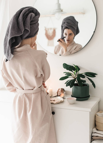 beautiful young woman in bathrobe and towel on her face and looking at camera while standing in bathroom - Foto, afbeelding