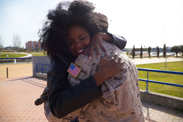 La mujer afroamericana y el soldado estadounidense que acaba de llegar de la guerra en una misión de paz se abrazan firmemente. La mujer es feliz. Concepto guerra y ejército, paz y misión. - Foto, imagen