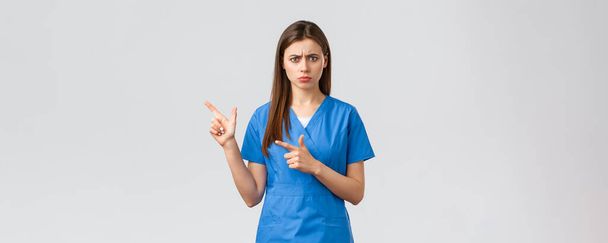 Healthcare workers, prevent virus, insurance and medicine concept. Concerned and alarmed young female doctor or nurse in blue scrubs frowning anxious, pointing fingers left at important info - Photo, Image
