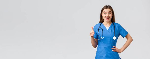 Медичні працівники, охорона здоров'я, коїд-19 та концепція вакцинації. Здивована збуджена медсестра або лікарка в блакитних скрабах зі стетоскопом, показують великі пальці на схвалення, як ідея, посміхаючись
 - Фото, зображення