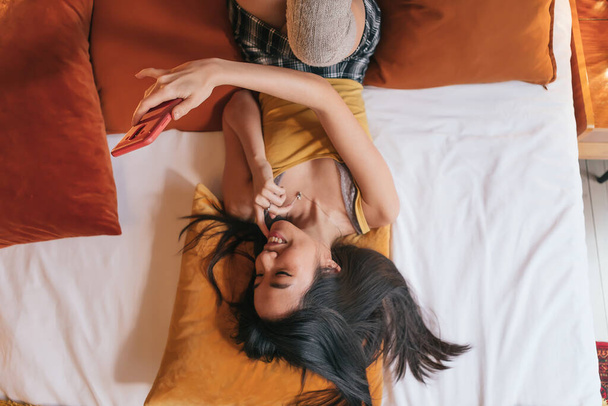 Draufsicht auf lächelnde asiatische junge Frau, die Handy im Bett hält und zusieht - Fokus auf Handy - Foto, Bild
