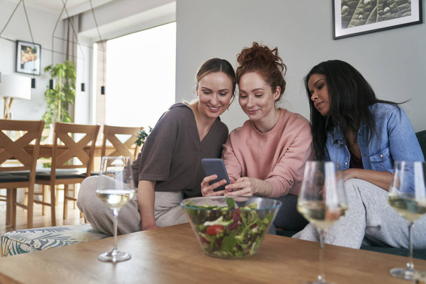 Trzech białych przyjaciół uśmiechniętych podczas przeglądania smartfona na kanapie       - Zdjęcie, obraz