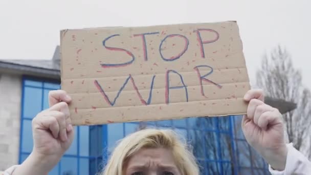 Украинка с светлыми волосами и серьезным жестом, с голубым и желтым украинским флагом с надписью: "Остановите войну", на улице, крича в знак протеста против войны между Украиной и Россией. - Кадры, видео