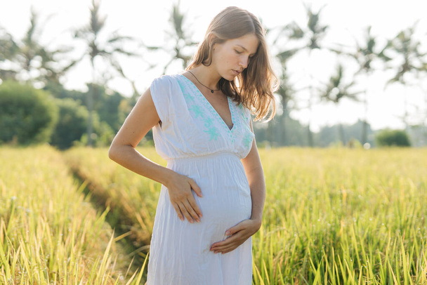 Pregnant woman stand at the field with green grass and embrace her belly. Sunshine summer vibes - Фото, зображення