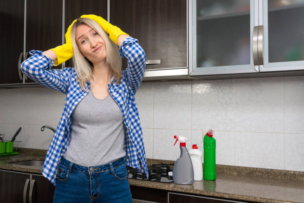 Mutfakta sarı eldivenli genç sarışın kadın başını tutuyor ve duygusal olarak yaklaşan temizlikten rahatsız oluyor ve tatmin olmuyor. - Fotoğraf, Görsel