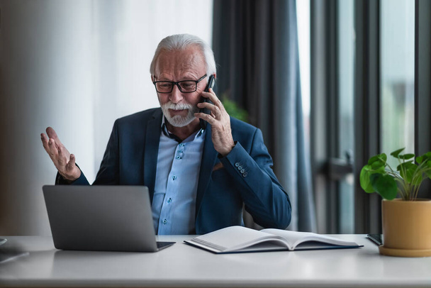 Portrait of mature senior businessman entrepreneur in casual business office making phone call and gesturing while working with laptop and examining, looking, going through data and reports. - Fotó, kép