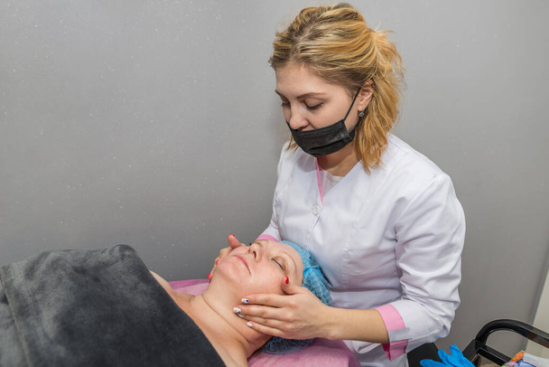An adult woman makes a facial massage at a beautician in a spa salon. Professional female cosmetologist makes facial massage to a client in a cosmetology room - Фото, зображення