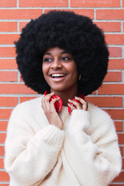Porträt einer fröhlich-charmanten verträumten afrikanisch-amerikanischen Frau mit weißem perfektem Lächeln, Afro-Frisur, weißem Pullover und roten Kopfhörern auf dem Hals im Freien beim Wegschauen. Backsteinmauer Hintergrund - Foto, Bild