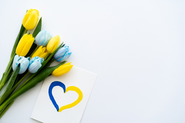 Draufsicht der Karte mit schmerzhaftem Herzsymbol in der Nähe gelber und blauer Tulpen auf weißem Hintergrund  - Foto, Bild