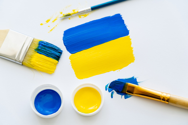 Vista dall'alto della bandiera ucraina dipinta vicino a vernici e pennelli su sfondo bianco  - Foto, immagini