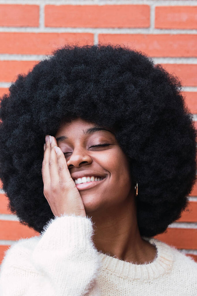 Portrait of cheerful african american woman with white perfect smile, long afro hair and white jumper covering eye against brick wall. Beautiful afro woman with eyes closed. Dentist concept - Foto, imagen