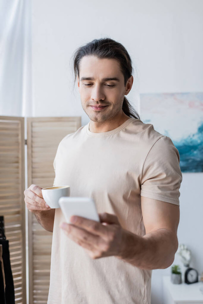 Mann im T-Shirt hält Tasse Kaffee und benutzt Smartphone - Foto, Bild