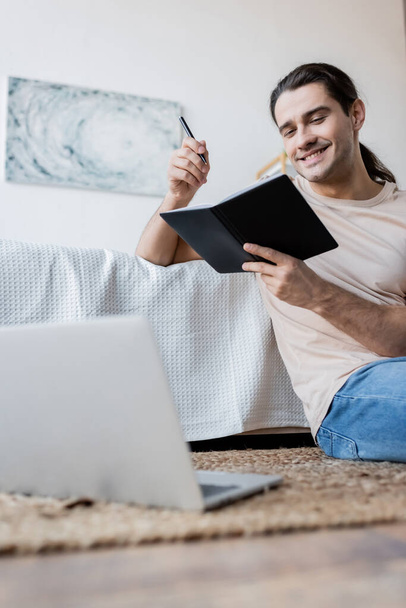 cheerful man with long hair holding notebook and pen near blurred laptop in bedroom  - Fotó, kép