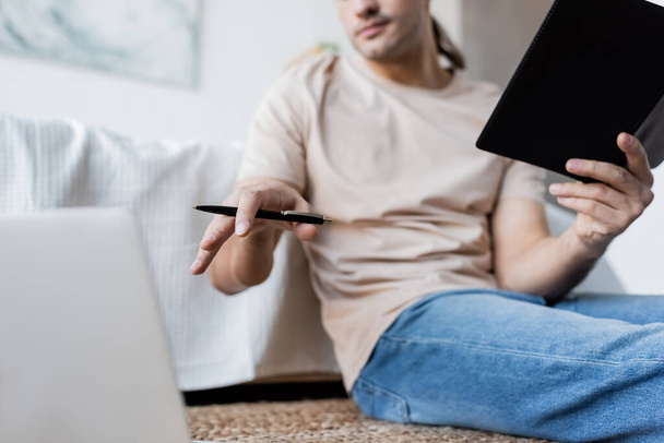cropped view of blurred man holding notebook and pen near laptop in bedroom  - Foto, afbeelding
