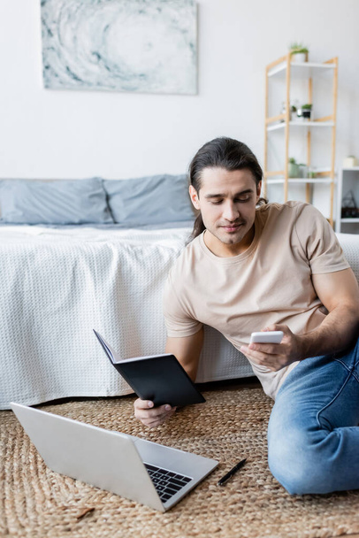 man holding notebook and smartphone near laptop in bedroom  - Fotó, kép
