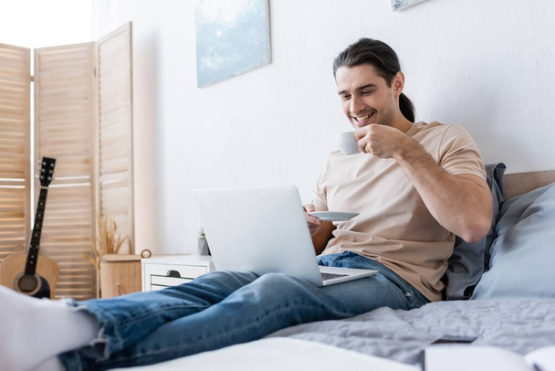 cheerful man with long hair holding cup of coffee while using laptop in bedroom - Valokuva, kuva