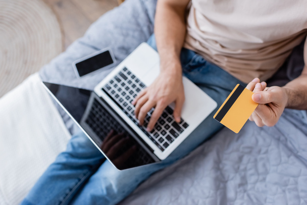 top view of man holding credit card and using laptop near smartphone on bed - Fotoğraf, Görsel