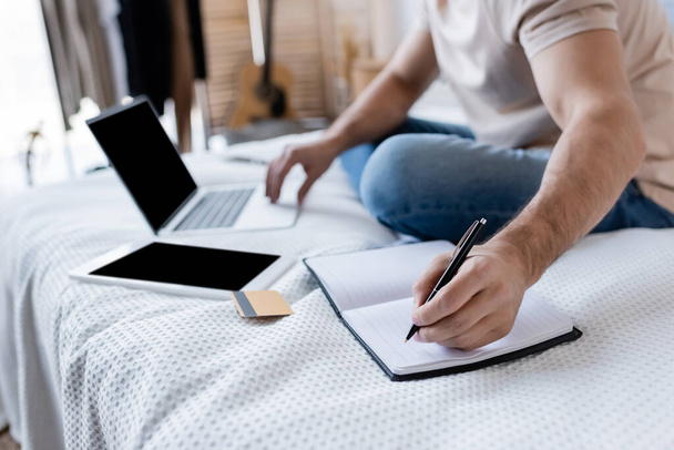 vista recortada del hombre escribiendo en el cuaderno y el uso de la computadora portátil cerca de la tarjeta de crédito en la cama - Foto, imagen