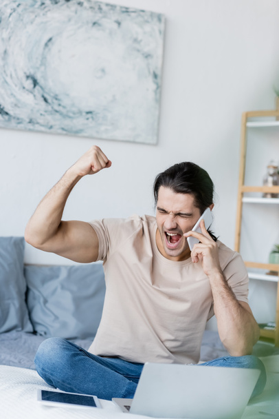 izgatott férfi beszél a mobiltelefon közelében laptop a hálószobában  - Fotó, kép