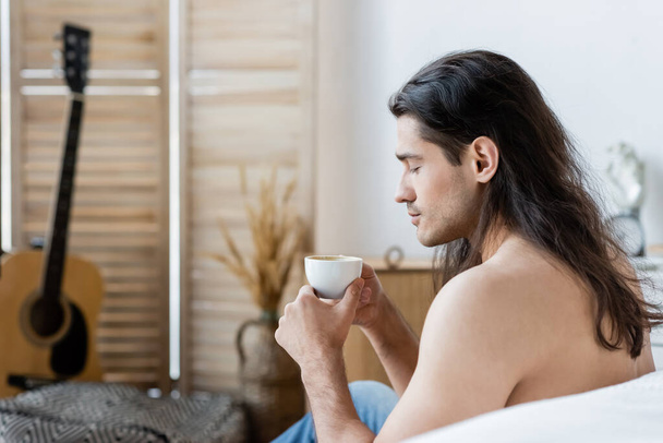 Seitenansicht eines hemdlosen Mannes mit langen Haaren, der eine Tasse Kaffee hält  - Foto, Bild