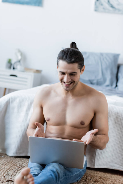 glücklicher Mann mit langen Haaren sitzt und Laptop in Bettnähe benutzt - Foto, Bild